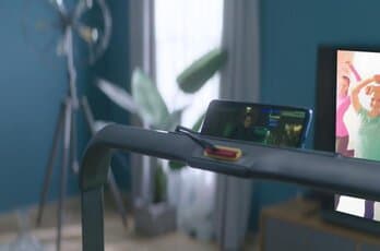 a phone secured on the handle of a treadmill with television running in the background